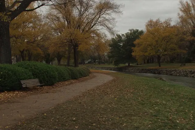 A riverside path through a city park in autumn, with a bench under yellow trees
