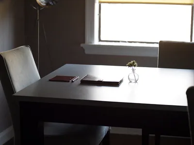 A dining table by a window with a closed leather folio, a pen and a small vase