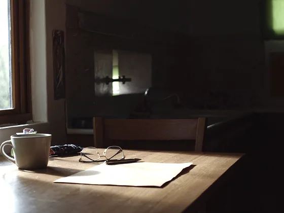 A kitchen table by a window with reading glasses, a pen, a blank sheet of paper and a mug